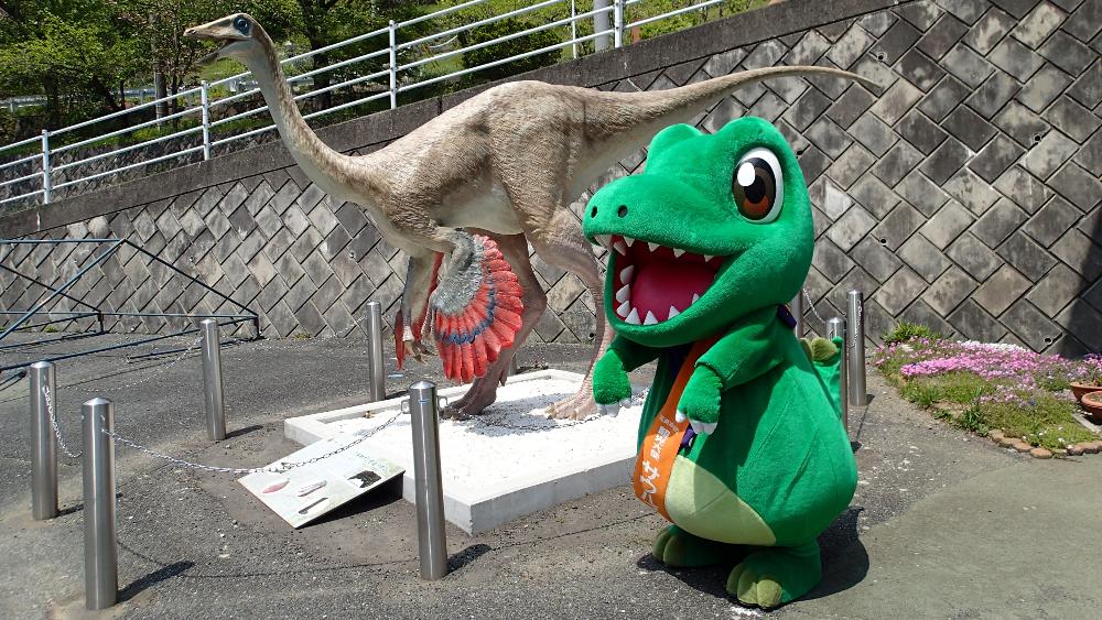 屋外に設置された恐竜のモニュメントの傍に立つ神流町観光大使サウルスくんの写真