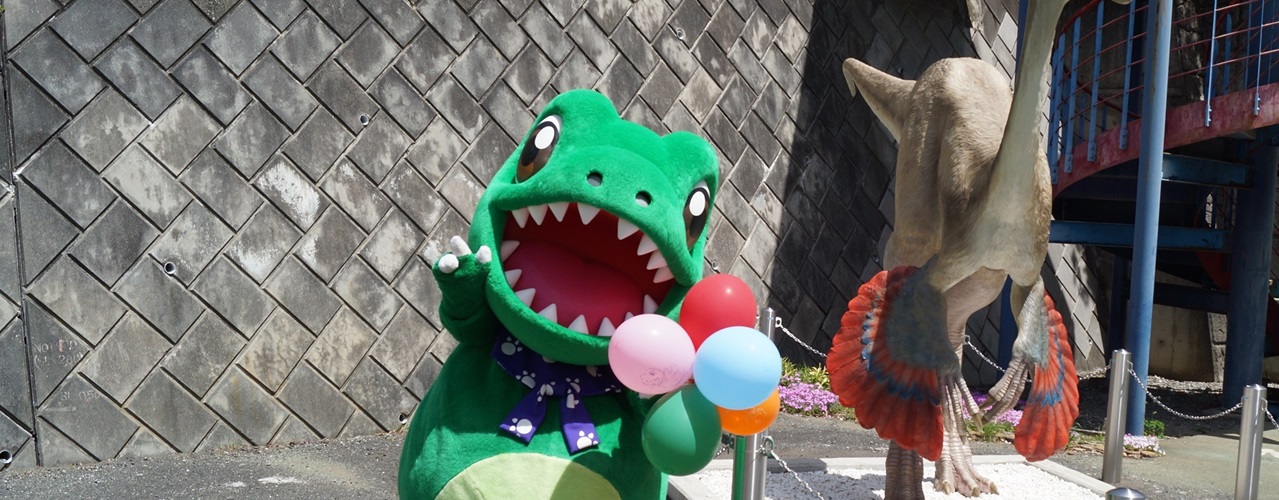 口をあんぐり開けた緑色の恐竜神流町観光大使サウルスが風船を持ってポーズを決めている様子。